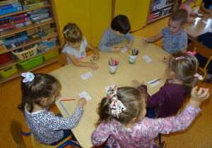 Sześcioro dzieci siedzi przy stoliku na którym rozłożone są kubeczki z kolorowymi kredkami. Przed każdym dzieckiem leży kartka z obrazkami kredek ułożonymi w różne kompozycje. Pięcioro dzieci sięga do kubeczków po kredki i próbują odtworzyć układ kredek z obrazka. Jedna z dziewczynek unosi rękę w gorę i oczekuje podejścia pani.